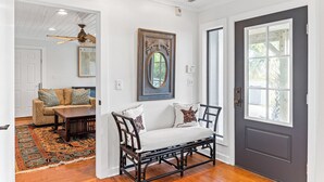 A hallway with a dark door, a white wall with a decorative mirror, and a patterned rug leading to a seating area with a bench and cushions.