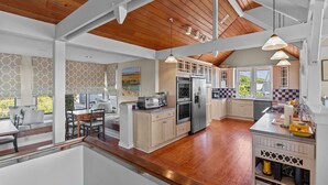 A spacious kitchen with wooden flooring, white cabinetry, and a large island. There are multiple windows providing natural light and views of the outdoors.