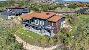 Ein modernes, zweistöckiges Haus mit großer Terrasse und Steinfundament, eingebettet in eine hügelige Landschaft.