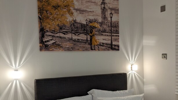 Une chambre à coucher avec un lit, du linge de lit blanc, une tête de lit sombre et un tableau représentant un pont et une tour de l’horloge.