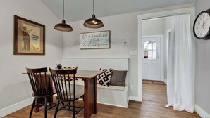 A dining area with a wooden table and chairs, a bench with cushions, pendant lights, a framed poster, and a wall clock.