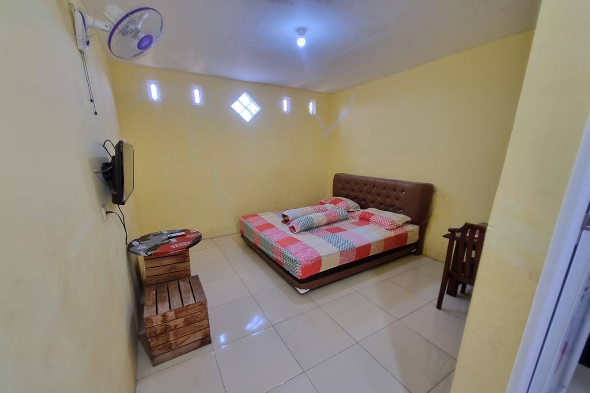 Une chambre à coucher avec un lit, un banc en bois, un ventilateur fixé au mur et un luminaire au plafond.