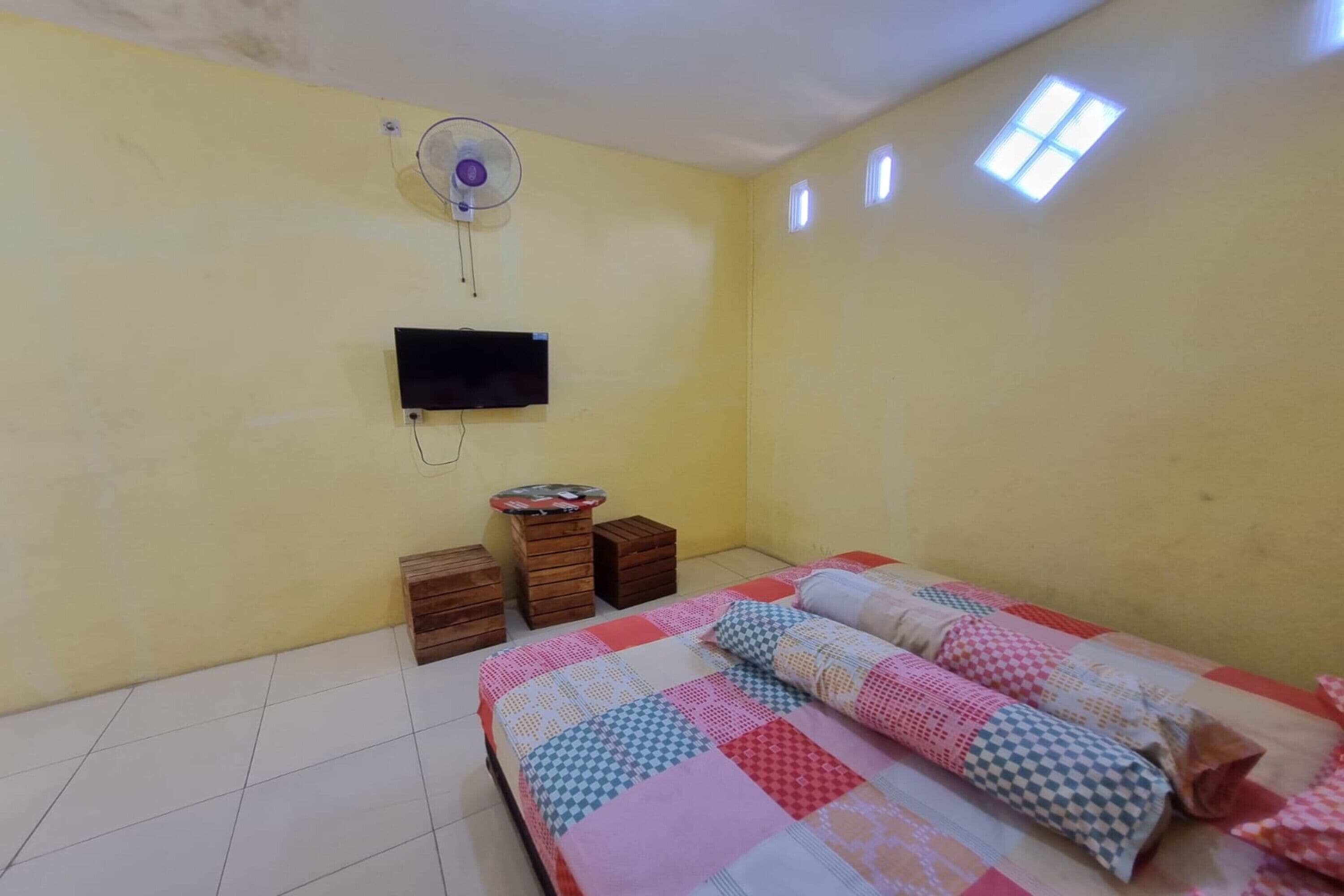 Una habitación con una cama, televisión montada en la pared, un ventilador y una mesita con cajones.