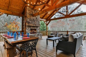 Une terrasse en bois avec une cheminée en pierre, un espace salon extérieur et un coin repas agrémenté de verres bleus et d’assiettes rouges.