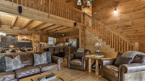 A spacious log cabin interior with a high ceiling, a kitchen area, and a cozy seating area with leather sofas and a coffee table.
