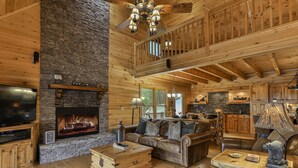 A log cabin interior with a stone fireplace, wooden furniture, a television, and a kitchen area.