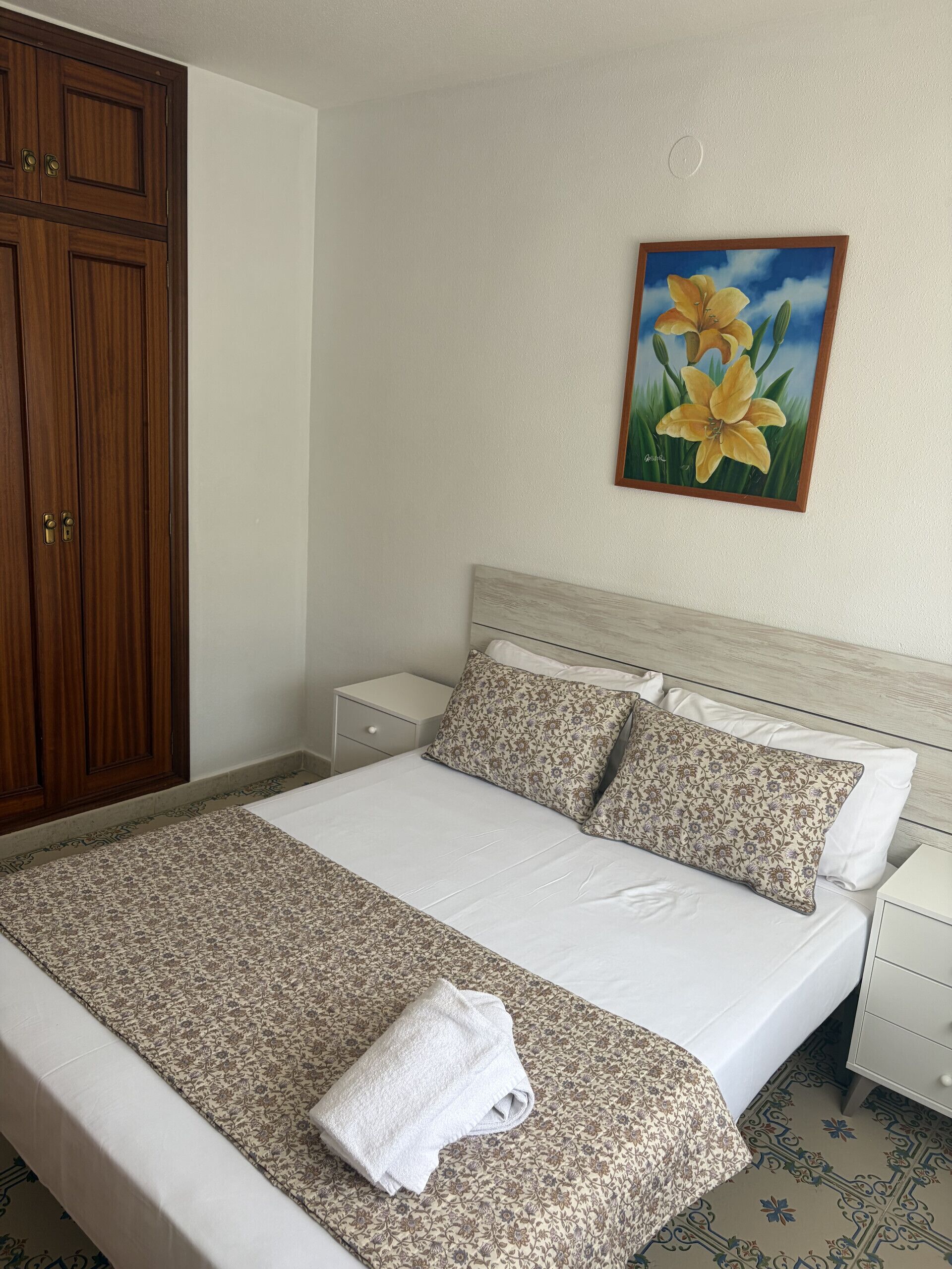 A neatly made bed with floral-patterned bedding, a white towel, and a painting of yellow flowers on the wall.