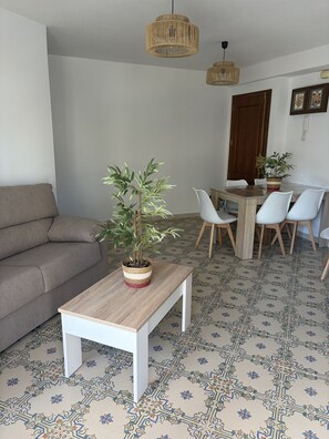 Una sala de estar con un sofá, una mesa de comedor con sillas, una planta en maceta y un lámpara colgante.
