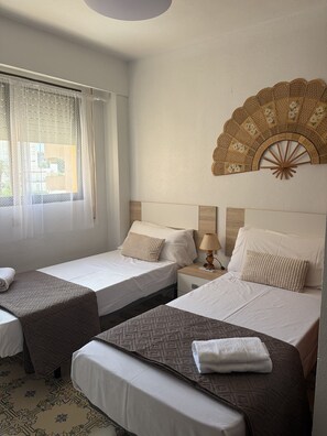 Una habitación de hotel con dos camas, una ventana con cortinas translúcidas, un abanico decorativo en la pared y un piso estampado.