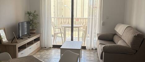A living room with a sofa, a dining table, chairs, a TV on a wooden cabinet, and a balcony with a view.
