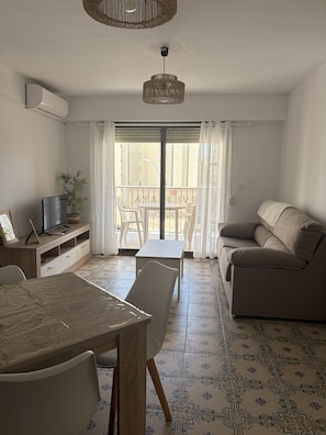 Una sala de estar con un sofá, una mesa de comedor, sillas, un televisor sobre un mueble de madera, y un balcón con vistas.