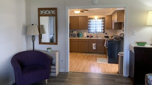 Un espacio habitable compacto con un sillón púrpura, una mesita auxiliar, un espejo y una kitchenette con armarios de madera, un microondas y un refrigerador.