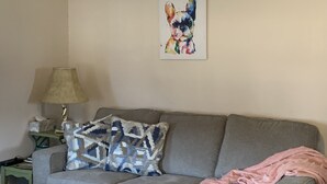 Una sala de estar con un sofá gris, cojines estampados, una lámpara y una obra de arte enmarcada en la pared.