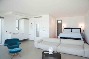 Moderni hotellihuone, jossa on sänky, sohva, tuoli, pieni pöytä ja kylpyhuone, joka näkyy avoimesta ovesta.