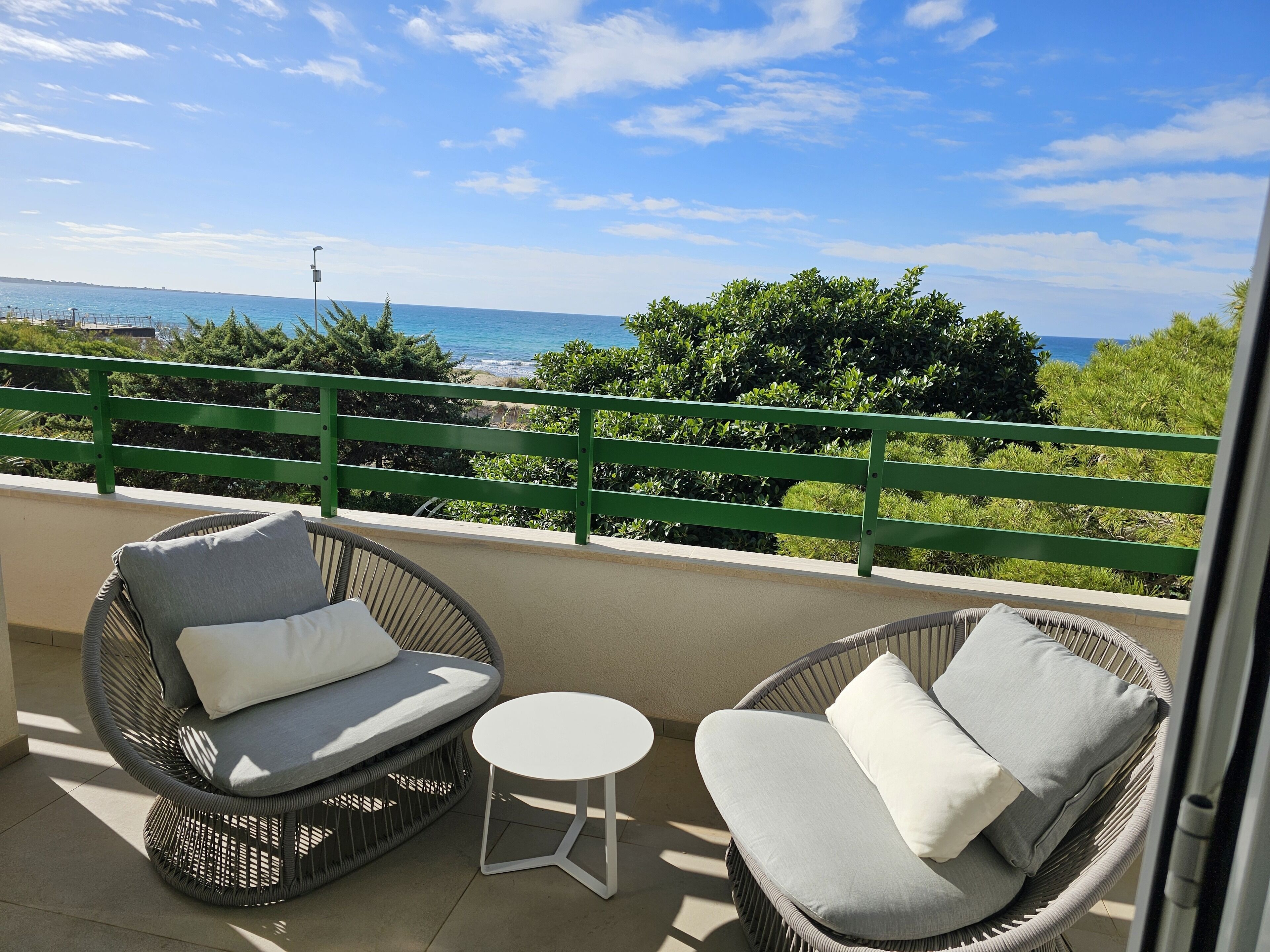 A balcony with wicker chairs and a small table, overlooking a coastal view with greenery and a clear sky.