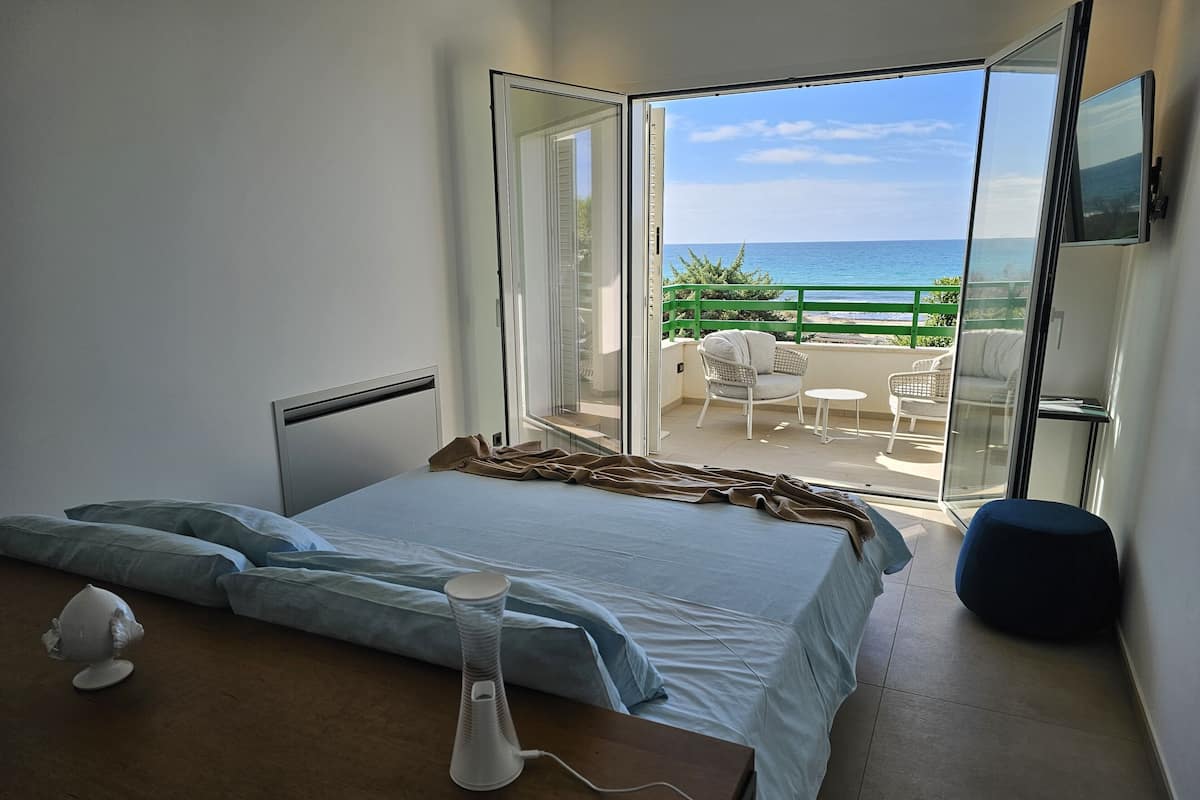 Une chambre à coucher avec un lit, une table de chevet avec une lampe, et une vue sur le balcon aménagé avec du mobilier d’extérieur.