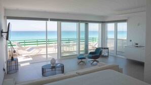 Un salon moderne doté d’un grand balcon donnant sur la mer, d’un canapé, de deux fauteuils, d’une petite table et d’un coin cuisine.