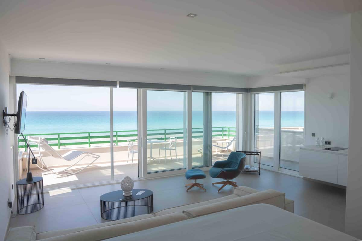 Un salon moderne doté d’un grand balcon donnant sur la mer, d’un canapé, de deux fauteuils, d’une petite table et d’un coin cuisine.