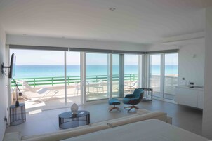 Una sala de estar moderna con un balcón grande con vista al mar, un sofá, dos sillones, una mesita y una zona de cocina.