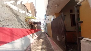 Une ruelle étroite, avec un mur rouge et blanc d’un côté et un mur jaune de l’autre. Une grille métallique se trouve sur le côté droit.