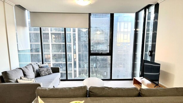 A modern living room with large windows, a grey sofa, a TV, and a plant.