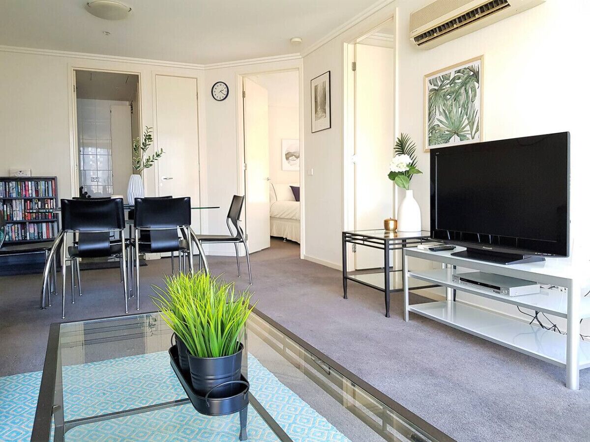 A living room with a flat-screen TV, a glass coffee table with a plant, two black chairs, a bookshelf, and a door leading to another room.