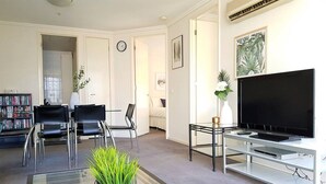 A living room with a flat-screen TV, a glass coffee table with a plant, two black chairs, a bookshelf, and a door leading to another room.