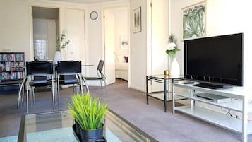 A living room with a flat-screen TV, a glass coffee table with a plant, two black chairs, a bookshelf, and a door leading to another room.
