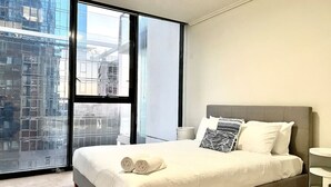 A modern hotel room with a large bed, a bedside table, and a view of a cityscape through floor-to-ceiling windows.
