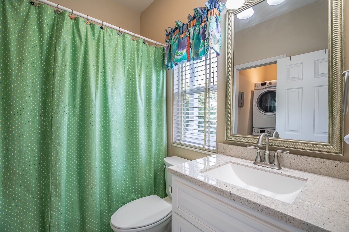 A bathroom with a green polka dot shower curtain, a white toilet, a sink with a mirror, and a window with blinds.