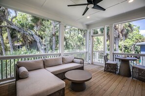 A covered outdoor area with a sofa, round table, and chairs, surrounded by wooden decking and large windows offering views of trees and a building. - New Construction, Close to Beach, 6 Seat Golf Cart Included - 713 Pompano Road (Fripp Island)
