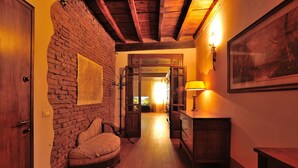 A room with a brick wall, wooden ceiling beams, a wooden floor, a round cushioned bench, a wooden cabinet, and a framed picture on the wall. - Orfeo's House (Modena)
