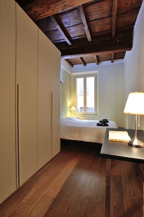 A bedroom with a wooden ceiling, a bed with a white comforter, a bedside table with a lamp, and a window with blinds.