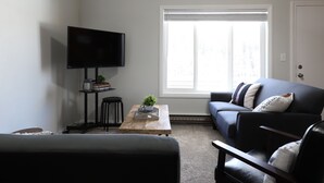 Una sala de estar moderna con un televisor de pantalla plana, una mesa de centro con libros y una planta, un sofá con cojines decorativos, y un ventanal que ofrece luz natural.