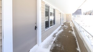 Un pasillo exterior cubierto con puerta corredera de vidrio, una terraza de madera y suelo cubierto de nieve.