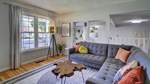 A living room with a grey sofa, a wooden coffee table, and a floor lamp. There is a window with curtains and a view of the outside.