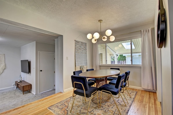 A dining area with a round table, six chairs, a chandelier, and a large window with curtains.