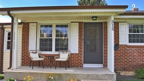 A two-story house with a brick exterior, a front porch with two chairs, and a mailbox.