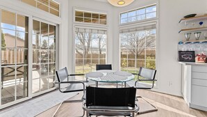 Um pátio iluminado pelo sol, com mesa de vidro e cadeiras de metal, janelas amplas e luminária no teto.