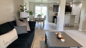 Ein Wohnzimmer mit dunkelgrauem Sofa, einem Couchtisch mit Dekoration, einem Essbereich mit Tisch und Stühlen sowie einer Küche mit Schränken und Arbeitsplatte.