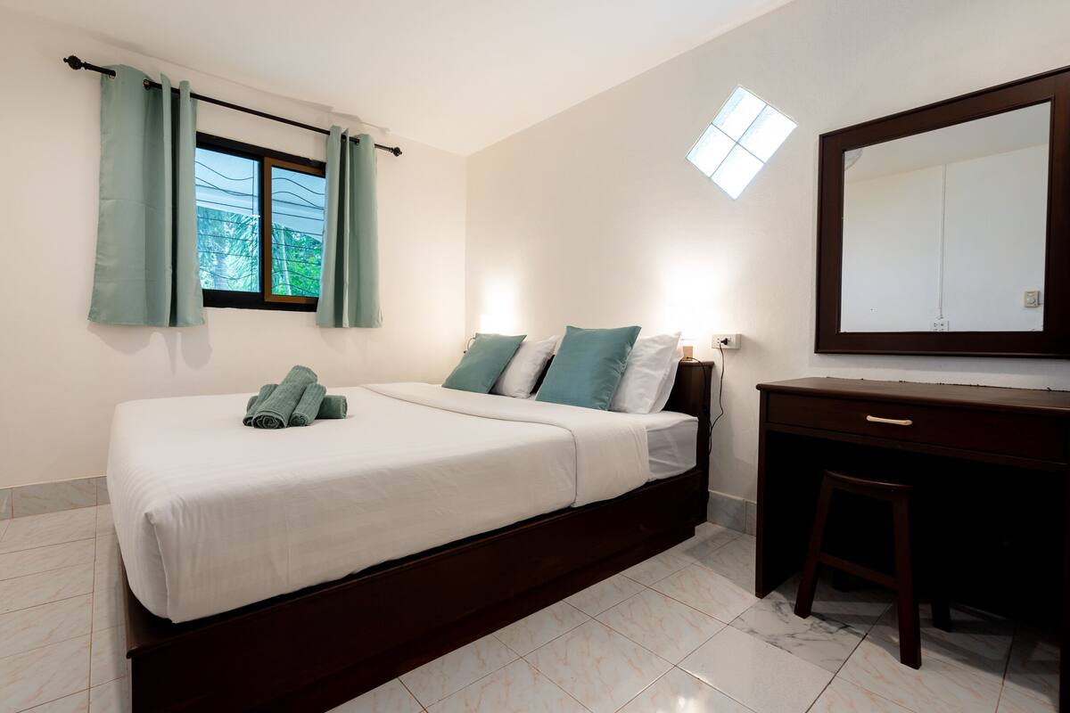 A bedroom with a bed, a desk, a chair, a window with curtains, a mirror, and a small skylight.