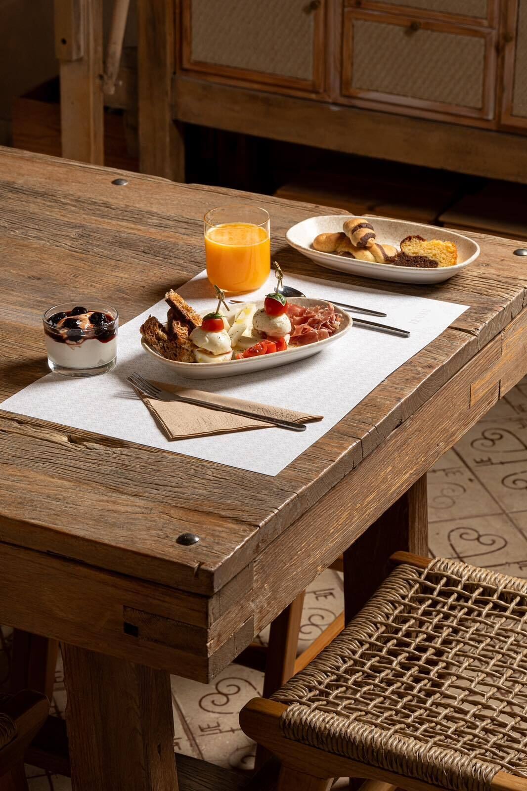 Una mesa de madera con una silla de mimbre, un plato de comida, un vaso de jugo de naranja y un tazón pequeño de yogur con bayas.