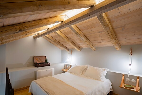 A bedroom with a sloped ceiling, a bed, bedside tables, and a small shelf with books. - ERAMAI (Patras)