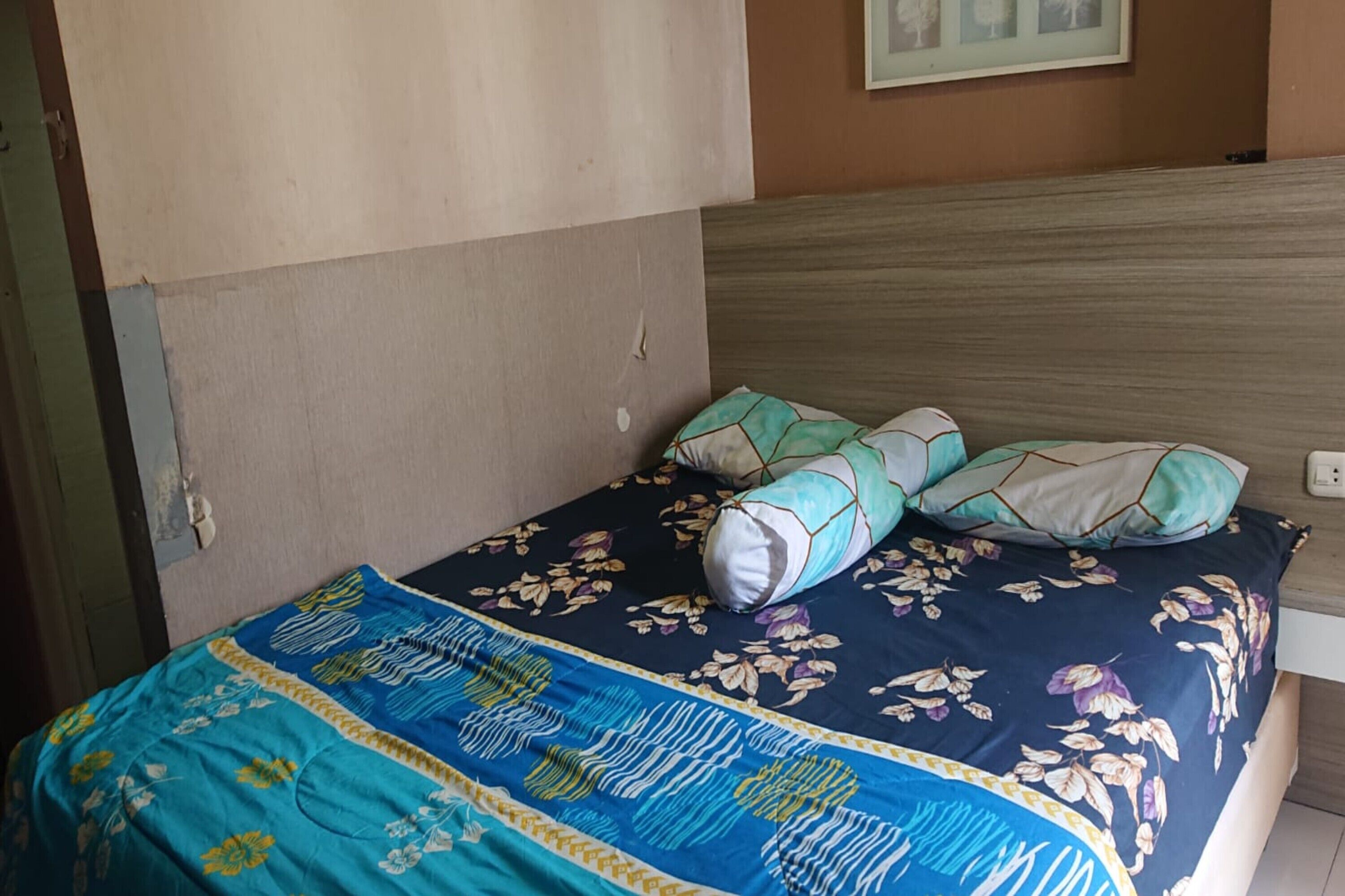 Una cama con colcha estampada, dos almohadas, cabecera de madera y una pared con un cuadro enmarcado.