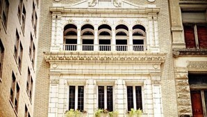 A building with a classical architectural style, featuring a central entrance with a sign reading "LYRIC PIANO CO." There are multiple windows with decorative moldings and a balcony with potted plants. - The Historic Lyric Penthouse with Private Rooftop (Cincinnati)