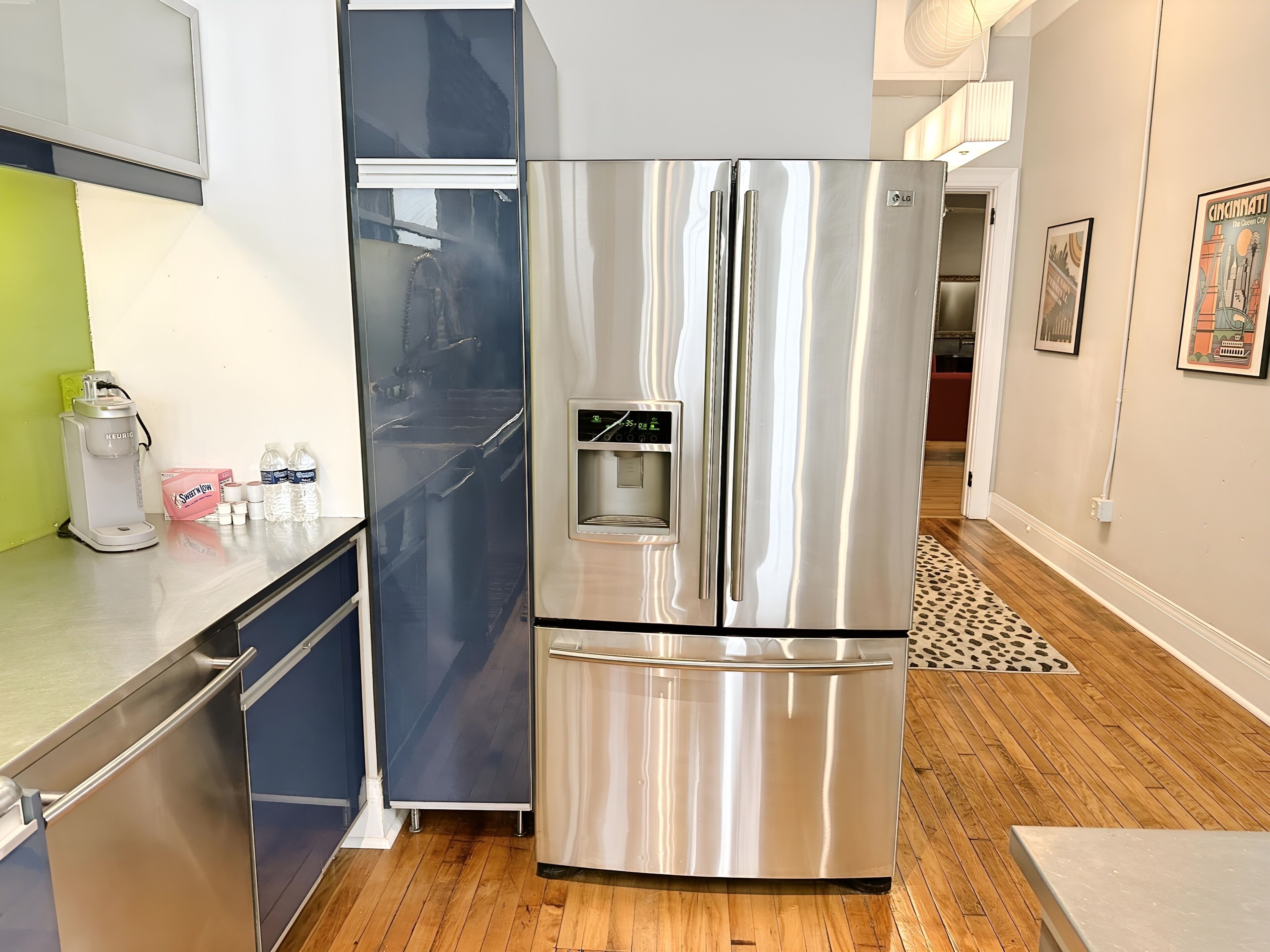 A stainless steel refrigerator with a water dispenser, a coffee maker, and a microwave on a wooden floor with a patterned rug.