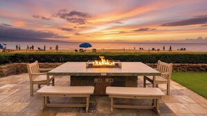 A patio with a fire feature, seating, and a view of the beach at sunset. - Whaler | Ocean View Studio Sleeps 4 | Car Incl. w/ 6+ Nights | WH2-661 by KBM (Lahaina)