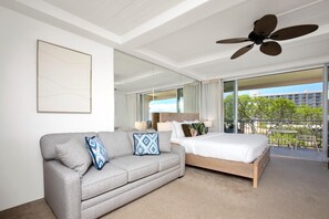 A modern hotel room with a grey sofa, a wooden bed, a ceiling fan, and a balcony with a view of trees and buildings.