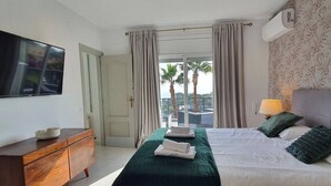 A hotel room with a bed, a TV, a dresser, and a balcony with a view of palm trees. - Opening Doors Premià Dalt Amazing views with pool (Premià de Dalt)