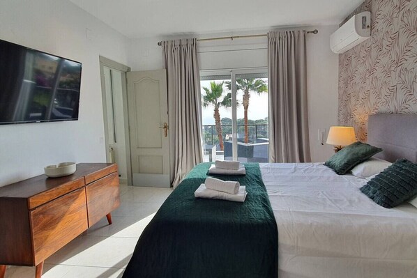 A hotel room with a bed, a TV, a dresser, and a balcony with a view of palm trees.